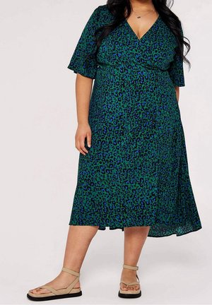 Mujer con vestido midi de estampado de leopardo verde y azul con escote en V y sandalias beige con tira en el tobillo, sobre un fondo liso.