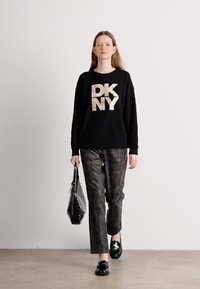 Svart hettegenser med metallisk gull "DKNY" logo, kombinert med mørke mønstrede bukser, svarte loafers og en svart håndveske med nagler.