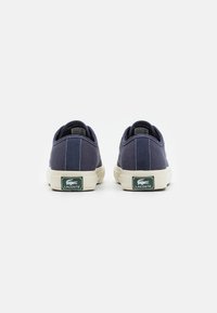 Tengerészkék vászon sportcipők fehér gumi talppal, zöld Lacoste logóval a saroknál és varrási részletekkel a felső részen.