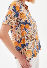 Short-sleeve, patterned top in beige with orange and blue floral designs. Lightweight fabric with a relaxed fit and subtle pleats at the front.