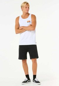 Weißes Tanktop mit "RIPCURL"-Logo und mehrfarbigen Streifen, kombiniert mit schwarzen Denim-Shorts und schwarzen Sneakers mit weißen Akzenten.