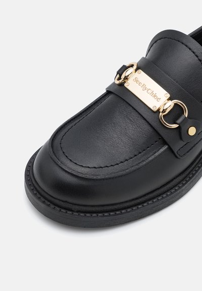 Mustad musta nahast loafer, millel on õmmeldud detailid ja kuldne metallplaat ning rõngasriist ülemise rihma peal, taustaks valge taust.
