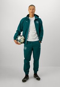 Giacca sportiva turchese e pantaloni in tessuto leggero con accenti di logo. Camicia bianca sotto e palla da calcio tenuta in mano. Scarpe da calcetto nere indossate.