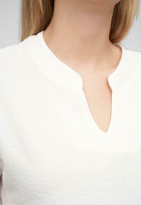 Hvid V-hals bluse med et blødt, struktureret stof, der har en subtil glans. Udsmykningen ved halsudskæringen har en lille slidsdetalje for ekstra interesse.