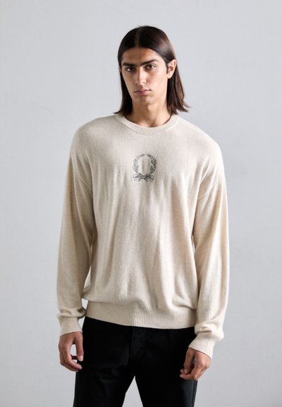 Jonge man met schouderlang donker haar, gekleed in een beige trui met een lauriertakembleem, staande voor een effen lichtgrijze achtergrond.