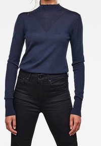 Top de punto azul marino con mangas largas y cuello alto. Presenta un panel triangular texturizado en el pecho. Combinado con jeans ajustados negros.