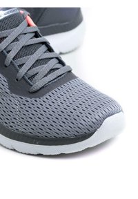 Chaussure de sport grise avec tige en maille, surface texturée, languette rembourrée et semelle blanche. Présente des lacets gris et des accents rouges près de la languette.
