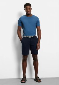 Pantaloncini blu navy abbinati a una t-shirt blu a maniche corte. Cintura con motivo intrecciato. L'outfit è completato da sandali neri.