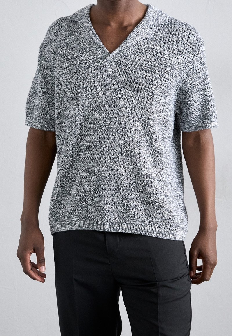 Homme portant un pull en maille gris à manches courtes avec un col en V et un pantalon noir sur un fond uni.