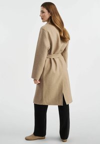 Beżowy płaszcz z wełny z paskiem, długimi rękawami i rozporkiem z tyłu, zestawiony z czarnymi spodniami i jasnymi butami.