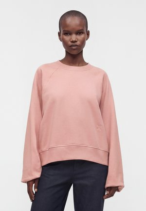 Person med kort hår iført en løstsiddende lyserød sweatshirt og mørke bukser, står mod en ensfarvet lys baggrund og vender fremad.