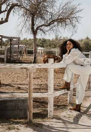 Femme vêtue de blanc assise sur une clôture en bois avec des chevaux pâturant dans une zone clôturée et un arbre sans feuilles à proximité.