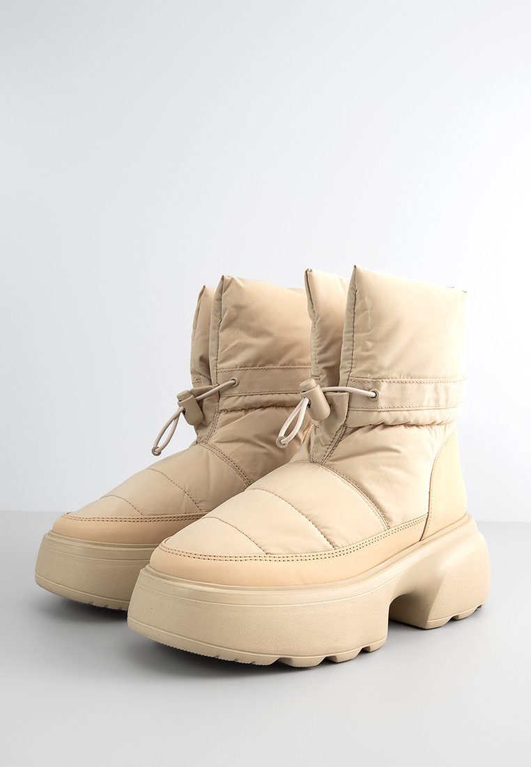 Beige Puff-Boots mit hohem Knöchel, gesteppter Oberseite und dicker Sohle. Mit Kordelzügen am Kragen und glatter synthetischer Textur.