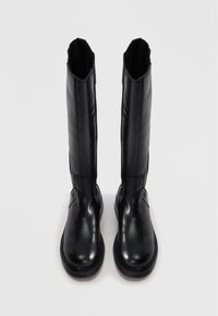 Une paire de grandes bottes en cuir noir avec des pointes arrondies et des semelles épaisses, se tenant debout sur une surface gris clair.