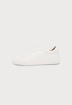 Sneaker bianca low-top con suola piatta, chiusura frontale con lacci e fodera interna marrone su sfondo grigio chiaro.