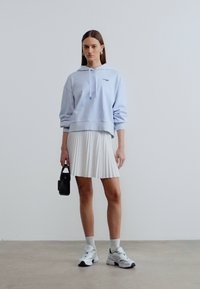 Femme portant un sweat à capuche bleu clair, jupe plissée blanche, baskets blanches, tenant un petit sac à main noir, debout sur un sol uni contre un mur blanc.
