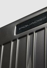 Graues Hartschalen-Koffersegment mit vertikalen Rippen. Verfügt über eine schwarze Metall-Logo-Plakette mit der weißen Aufschrift "PIQUADRO".