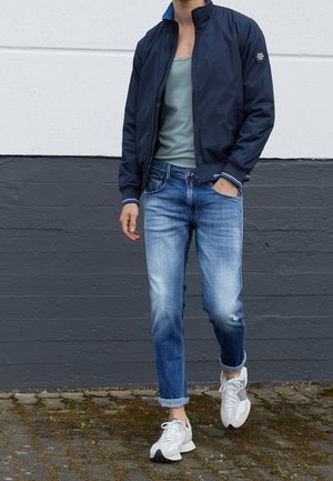 Person iført en navyjakke, grå tanktop, falmede blå jeans med opkørte ankler og hvide sneakers, går på en brostensbelagt fortov.