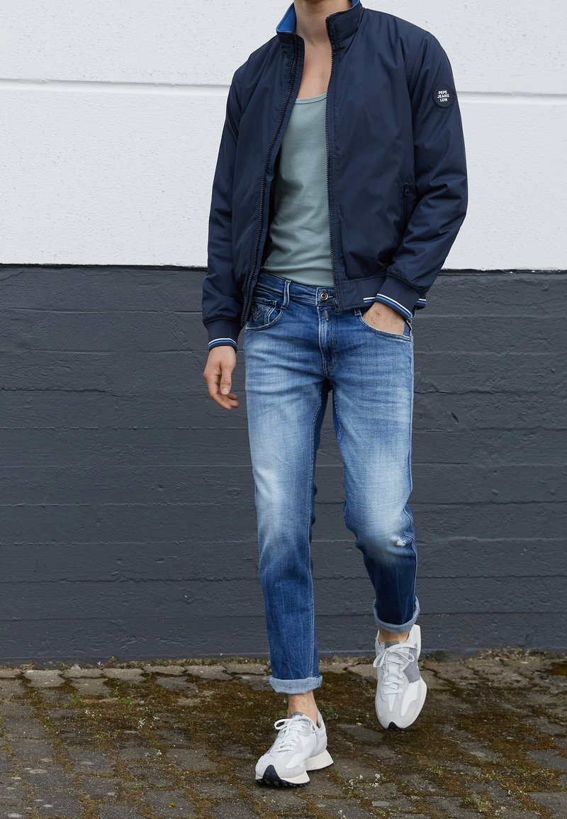 Personne portant une veste bleu marine, un débardeur gris, un jean bleu délavé retroussé aux chevilles, et des baskets blanches, marchant sur un pavé en pierre.