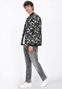 Camouflage bomberjack in groen en grijs, zwarte ribbels op de mouwen, gecombineerd met grijze jeans en witte sneakers. Model staat, kijkt naar links.