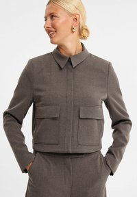 Brun cropped jakke lavet af tekstureret stof, med krave, lukning foran med knapper og to store klaplommer på brystet.