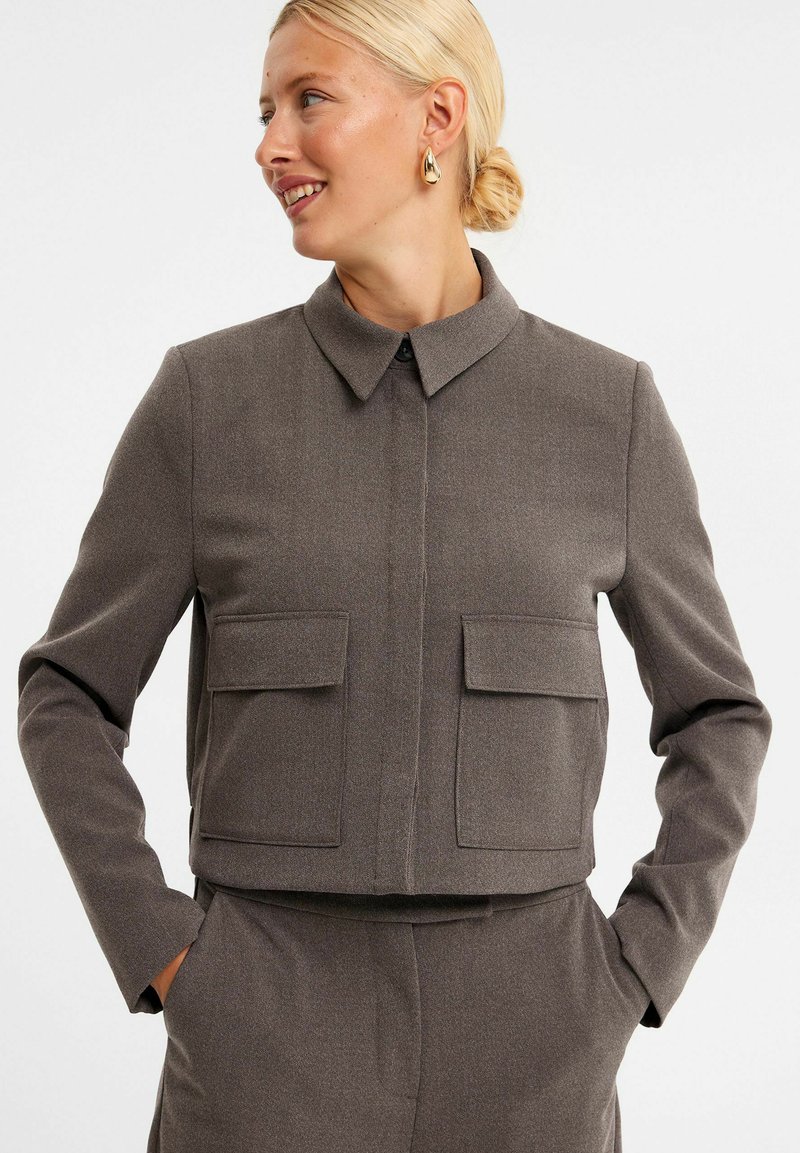 Brun cropped jakke lavet af tekstureret stof, med krave, lukning foran med knapper og to store klaplommer på brystet.