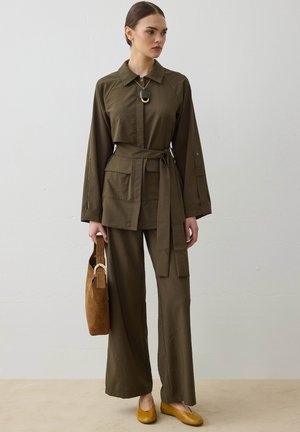 Frau trägt olivgrüne, taillierte Jacke und weit geschnittene Hose, hält eine braune Wildledertasche, dazu senfgelbe Ballerinas und eine Anhängerkette.