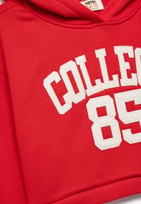 Punainen lyhyt huppari, jossa on valkoista "COLLEGE 85" -kirjontaa. Sileä pinta, säädettävä huppu ja ribbikuvioitu helma. Rentoa urheiluvaatemuotoilua.