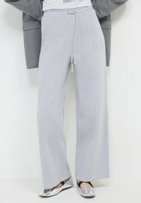 Pantaloni della tuta grigio chiaro con vestibilità rilassata, dotati di vita con coulisse, cuciture laterali e texture morbida. Abbinati a ballerine argentate.