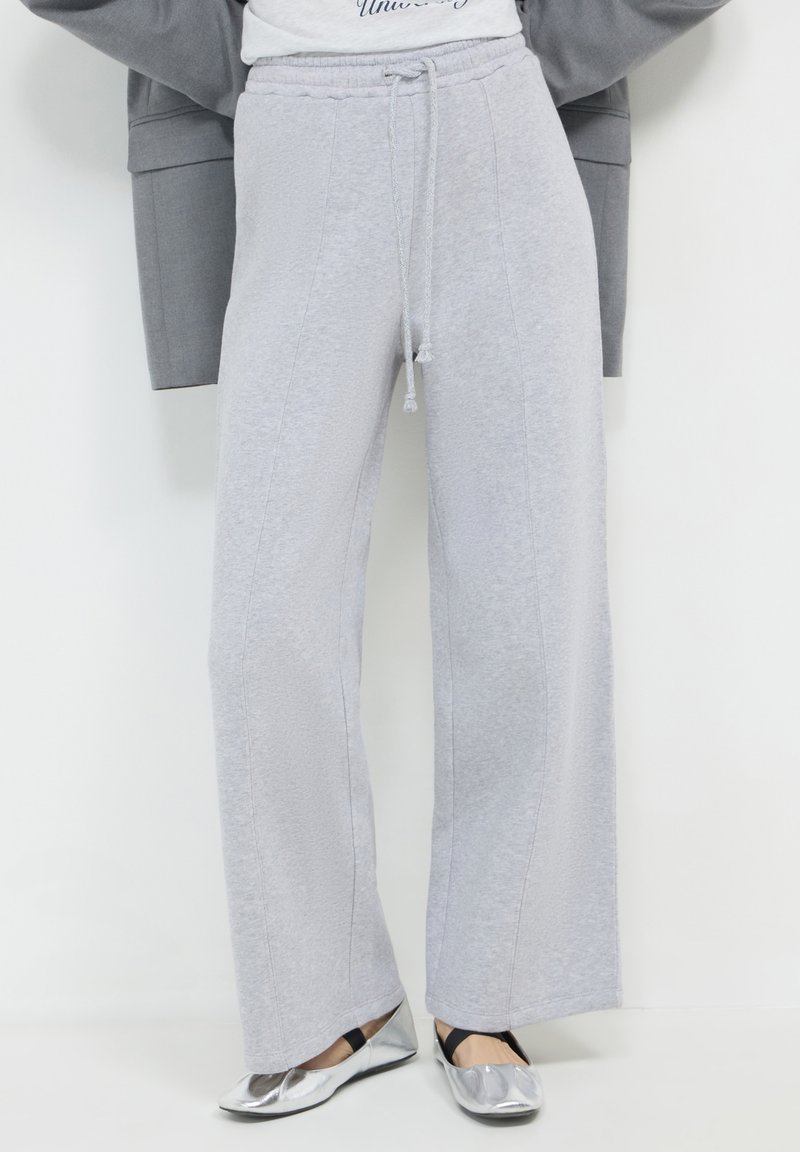 Pantaloni della tuta grigio chiaro con vestibilità rilassata, dotati di vita con coulisse, cuciture laterali e texture morbida. Abbinati a ballerine argentate.