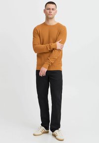 Brązowy sweter o fakturze z okrągłym dekoltem, w połączeniu z ciemnymi zwężającymi się spodniami i białymi sneakersami z brązowymi paskami po bokach.