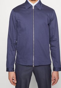 Mörkblå zip-jacka i mjukt tyg med spetsig krage, framfickor och detaljer i metallisk dragkedja.