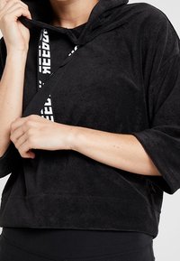 Sweat à capuche court noir en tissu doux, avec une capuche et des manches amples. Comprend une sangle de marque avec un texte blanc sur le devant.