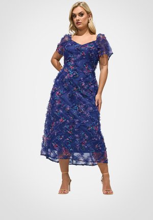 Kobieta w granatowej sukience midi w kwiaty z przezroczystymi krótkimi rękawami i beżowymi sandałami na wysokim obcasie, pozująca na jednolitym tle.