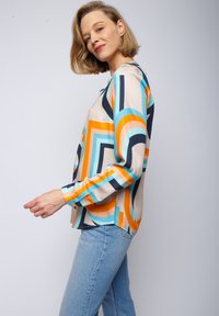 Kleurige blouse met geometrisch patroon in een zijden stof, met blauwe, oranje en zwarte lijnen, gecombineerd met lichtblauwe jeans. Lange mouwen, relaxte pasvorm.
