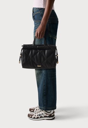 Black quilted handbag with two top handles, featuring gold accents and side drawstrings; paired with relaxed blue jeans and brown athletic shoes.