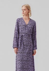 Lila blommig wrap-klänning med långa ärmar, featuring en V-hals, knuten midja och ett subtilt texturerat mönster mot en mörk bakgrund.