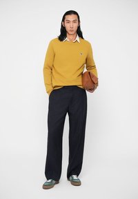 Kollane sviiter valge kraega, meresinised triibulised püksid ja rohelised tossud. Hoides pruuni kotti. Lihtne disain ja tekstuuriga materjalid.