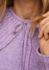 Donna che indossa un cardigan lavorato a maglia lavanda con un colletto decorato, abbottonato in cima, mentre tiene una ciocca di capelli biondi vicino al viso.