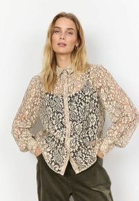 Lace blouse featuring a sheer design in beige and black, with floral patterns, buttoned front, and long sleeves. Textured, lightweight fabric.