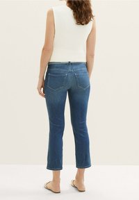 Mujer de pie, de espaldas, vistiendo unos vaqueros cortos azul, una camiseta sin mangas de punto blanco, y sandalias de lona beige, sobre un fondo claro.