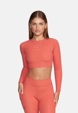 Kobieta w koralowym, długim, krótkim topie sportowym i legginsach z wysokim stanem, stojąca z jedną ręką na biodrze na jednolitym tle.