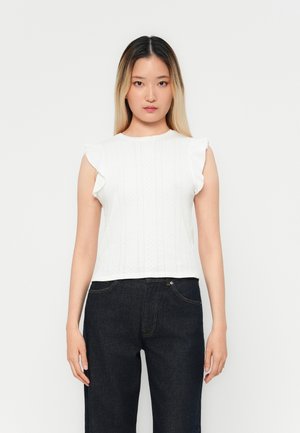 Haut blanc à manches courtes avec des épaules froncées, présentant un motif texturé. Porté avec un pantalon en denim foncé, sur un fond uni.