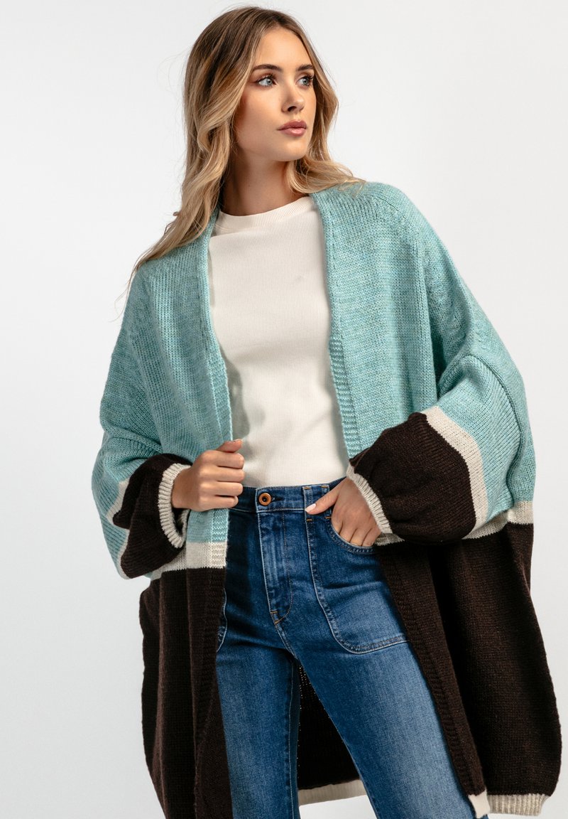 Przesadzony sweter dzianinowy w jasnoniebieskim, brązowym i kremowym kolorze. Posiada szerokie rękawy, otwarty przód i teksturowany materiał. Zestawiony z niebieskimi dżinsami.