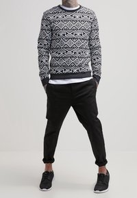 Person som bär en svart och vit geometriskt mönstrad tröja, svarta uppvikta byxor, svarta sneakers, står med händerna i fickorna.