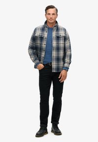 Camicia a quadri con tonalità blu e grigie, jeans neri e stivali neri. La stoffa ha una texture morbida, con due tasche sul petto e chiusure a bottoni.