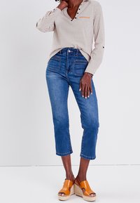 Blouse beige à motifs avec boutons noirs, associée à un jean bleu court et des sandales compensées orange. Tissu lisse et coupe structurée.