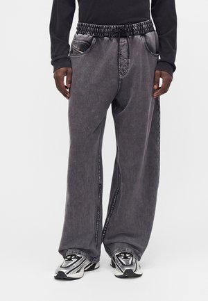 Person iført løse grå vaskede jeans med elastisk taljebånd og sølv-sorte-hvide sneakers, sort langærmet top, stående.