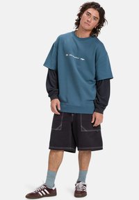 Sudadera de gran tamaño en color verde azulado con un gráfico blanco, superpuesta a una camiseta de manga larga negra. Pantalones cortos negros con costuras blancas y bolsillos. Zapatillas marrones.