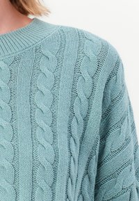 Lyseblå strikkegenser med kabelstrikk mønstre, ribbestrikket rund hals og teksturerte detaljer i stoffet.